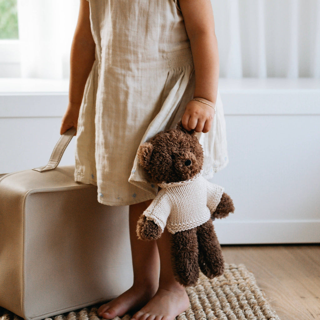 Child holding Milo Bear soft toy by the arm while standing barefoot, highlighting plush texture and knitted jumper detail.