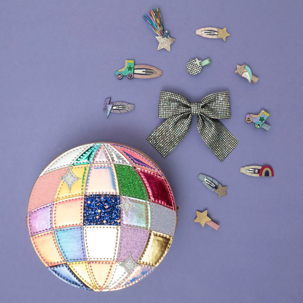 Flat lay of Mimi & Lula Disco Diva Clips and a patchwork disco ball-shaped bag with glitter and metallic details on a purple background.