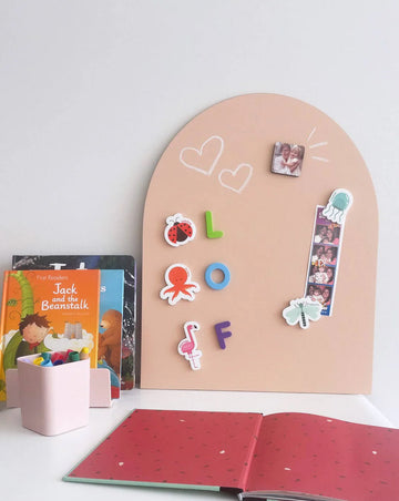 Mini MagPlay magnetic wall decal in a soft blush tone, styled with magnetic letters, photos, and stickers; surrounded by books, a pencil cup, and an open notebook on a child’s desk.