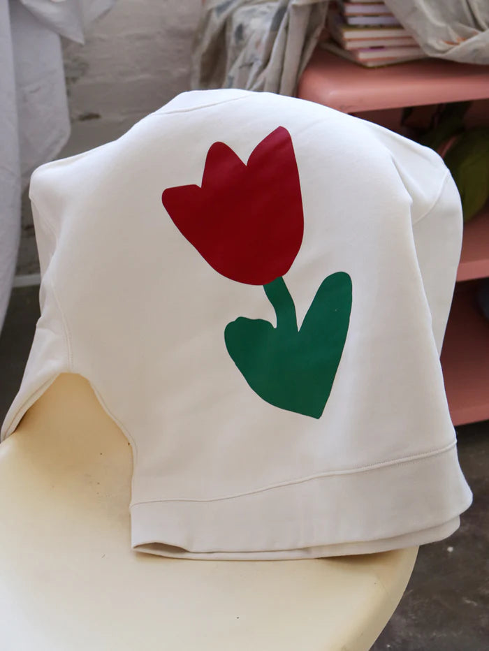 Mosey Me Tulip Sweater in Ecru draped over the back of a chair, showcasing the bold red tulip and green leaves on the soft cream fabric.