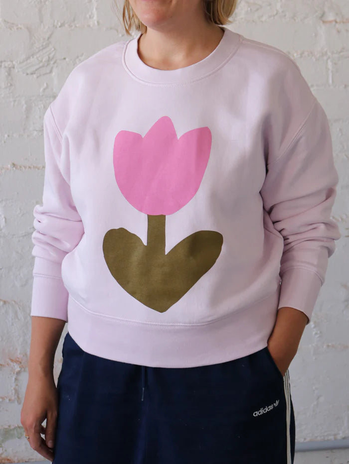 Front view of a model wearing the Mosey Me Tulip Sweater in Orchid, showing the oversized fit, drop shoulders, and bold tulip print on the chest, styled casually with navy pants.