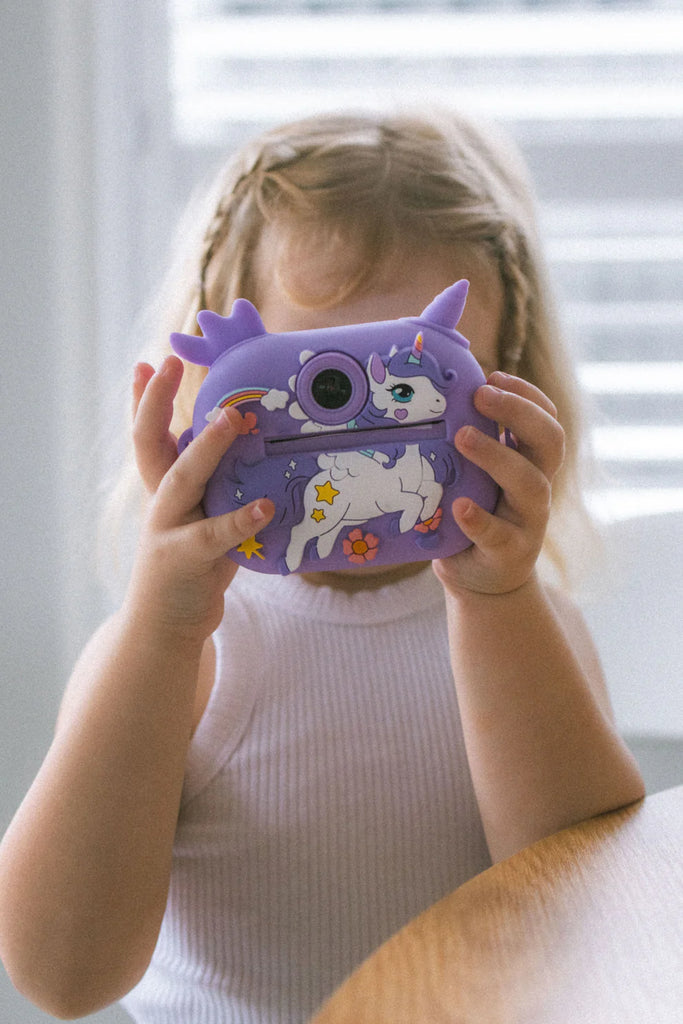 Little girl holding a purple unicorn instant print camera, smiling while pretending to take a photo indoors.