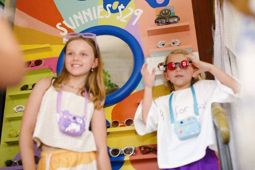 Two children wearing colorful instant print cameras around their necks while playing in a fun sunglasses shop.