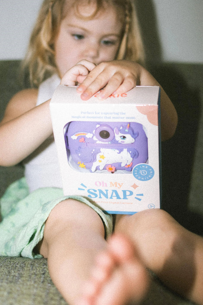 Child holding a boxed purple unicorn instant print camera, excited to open her new toy camera.