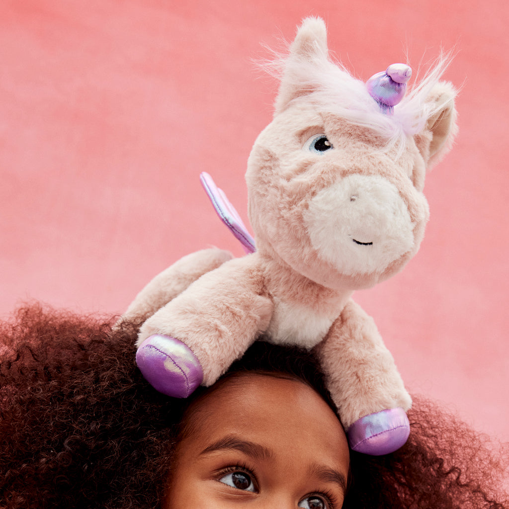 Shimmer unicorn plush balanced on a girl’s head – Playful moment with posable unicorn showing lavender horn and purple wings.