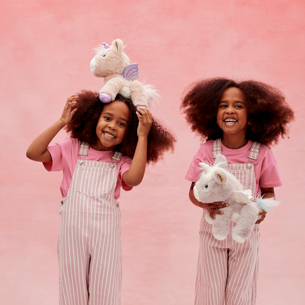 Two children playing with Shimmer the Dinkum Unicorn – Girls laughing and holding the plush unicorn against a pink background.