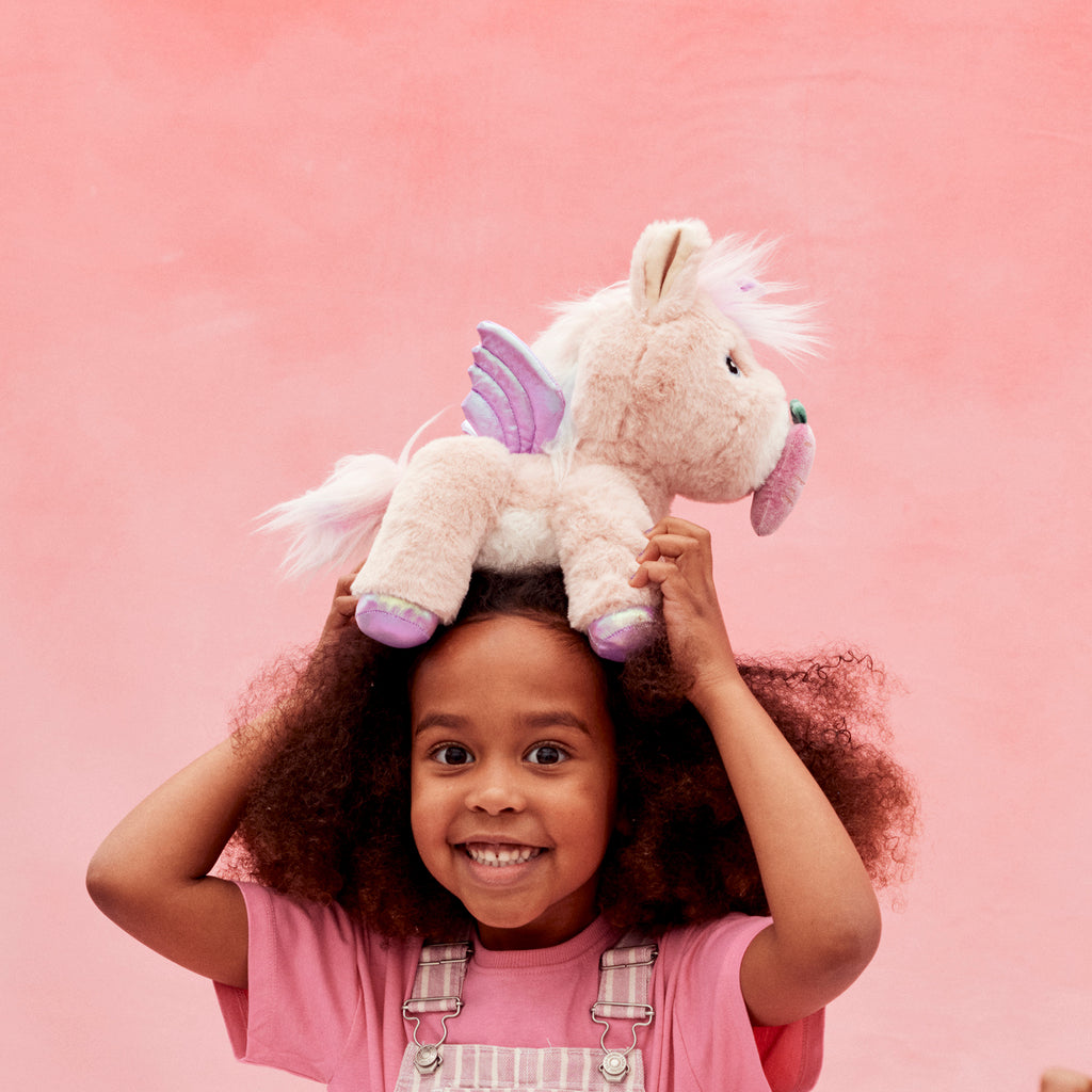 Close-up of smiling girl holding Shimmer unicorn on her head – Child with curly hair balancing pink unicorn plush with white mane.