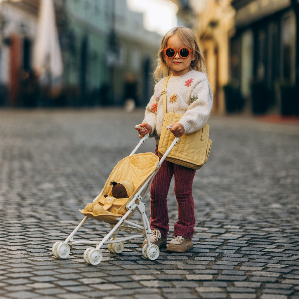 Toddler pushing Dinkum Doll in honey stroller outdoors – Child strolling doll along cobblestone street in honey-colored stroller.