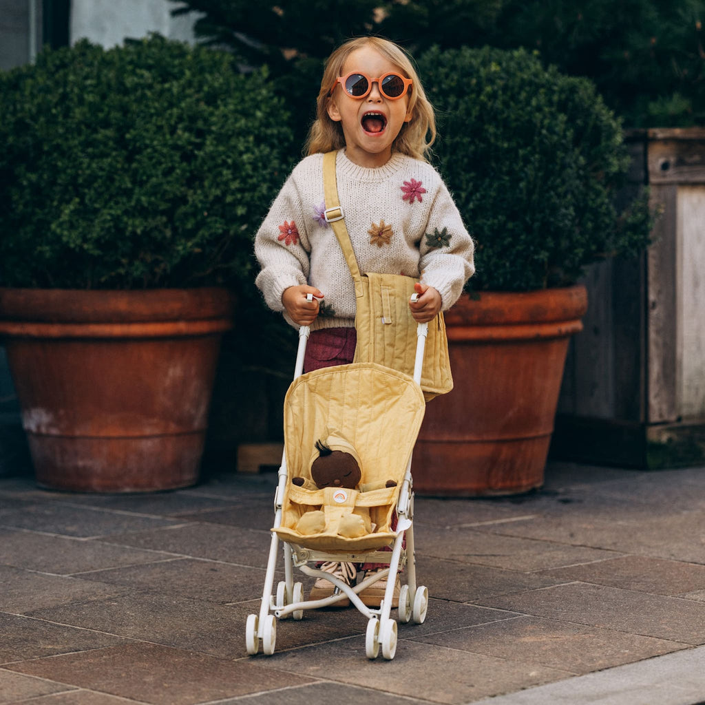 Child smiling while holding honey stroller outdoors – Toddler happily walking with doll pram in honey fabric seat design.