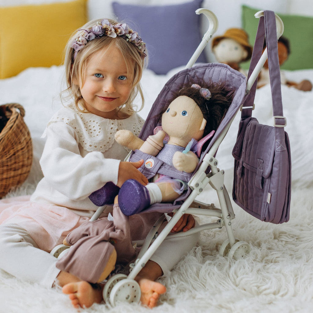 Child smiling beside Dinkum Doll in stroller indoors – Girl seated next to lavender stroller with doll, enjoying playtime.