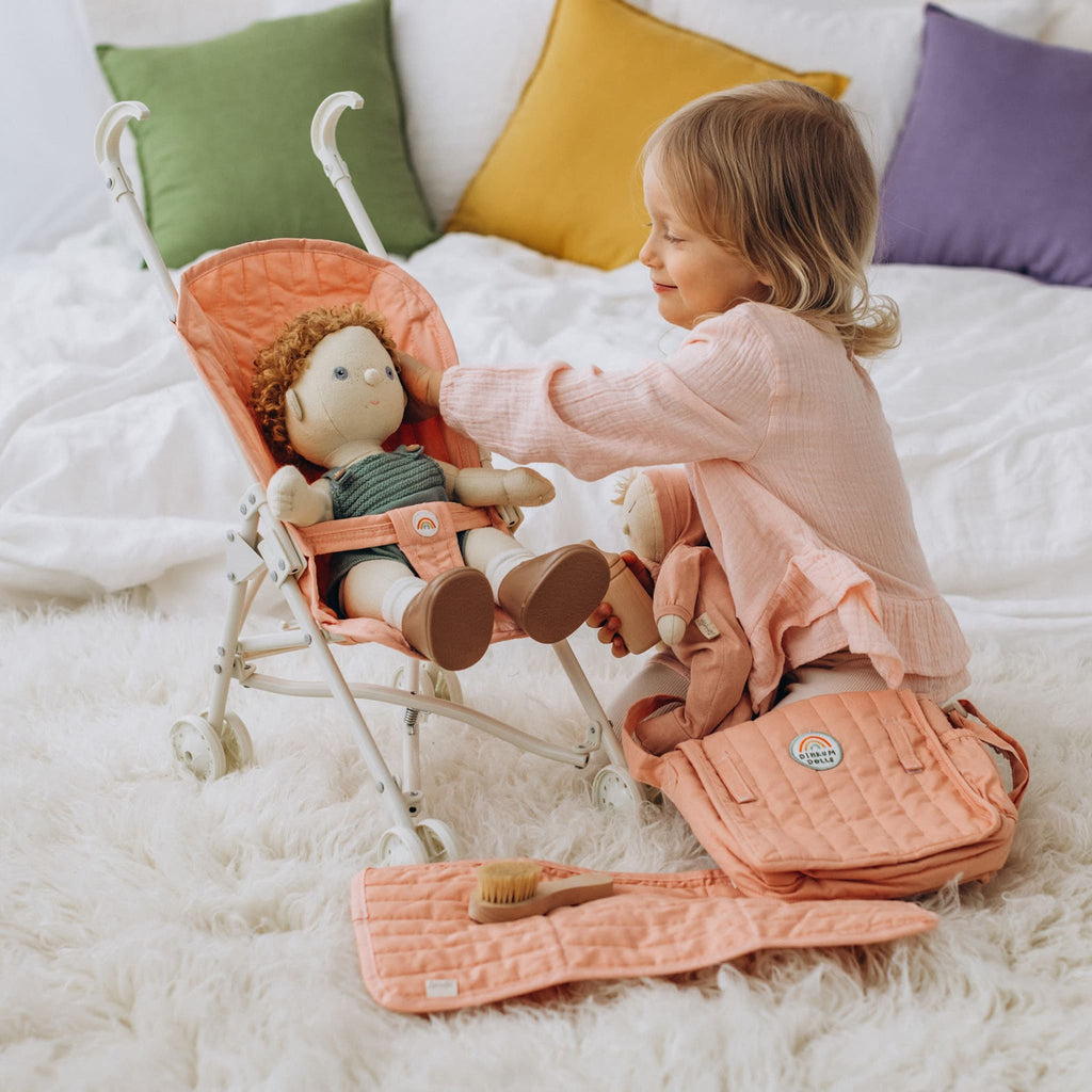 Toddler kneeling beside stroller indoors – Child arranging doll in rose stroller, quilted fabric and metal frame visible.