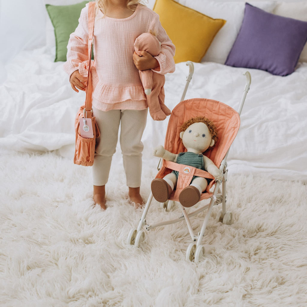 Child standing with stroller and doll indoors – Doll seated in stroller while child holds matching carry bag.