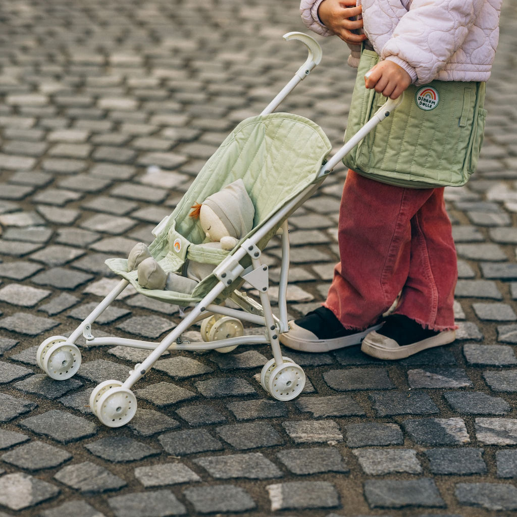 Toddler walking outdoors with Sollie Stroller Sage – Child strolling on cobblestone street with doll pram in sage green.