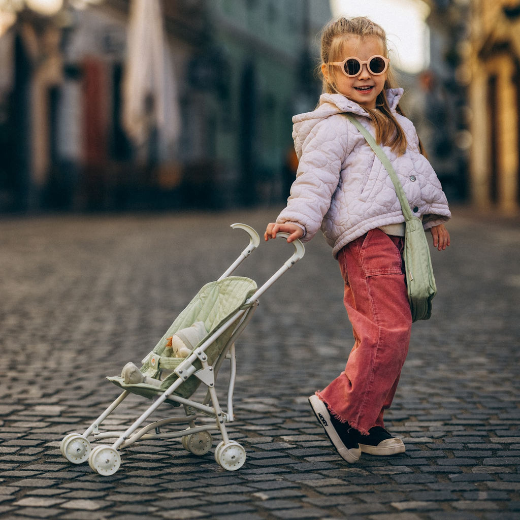 Toddler walking outdoors with Sollie Stroller Sage – Child strolling on cobblestone street with doll pram in sage green.