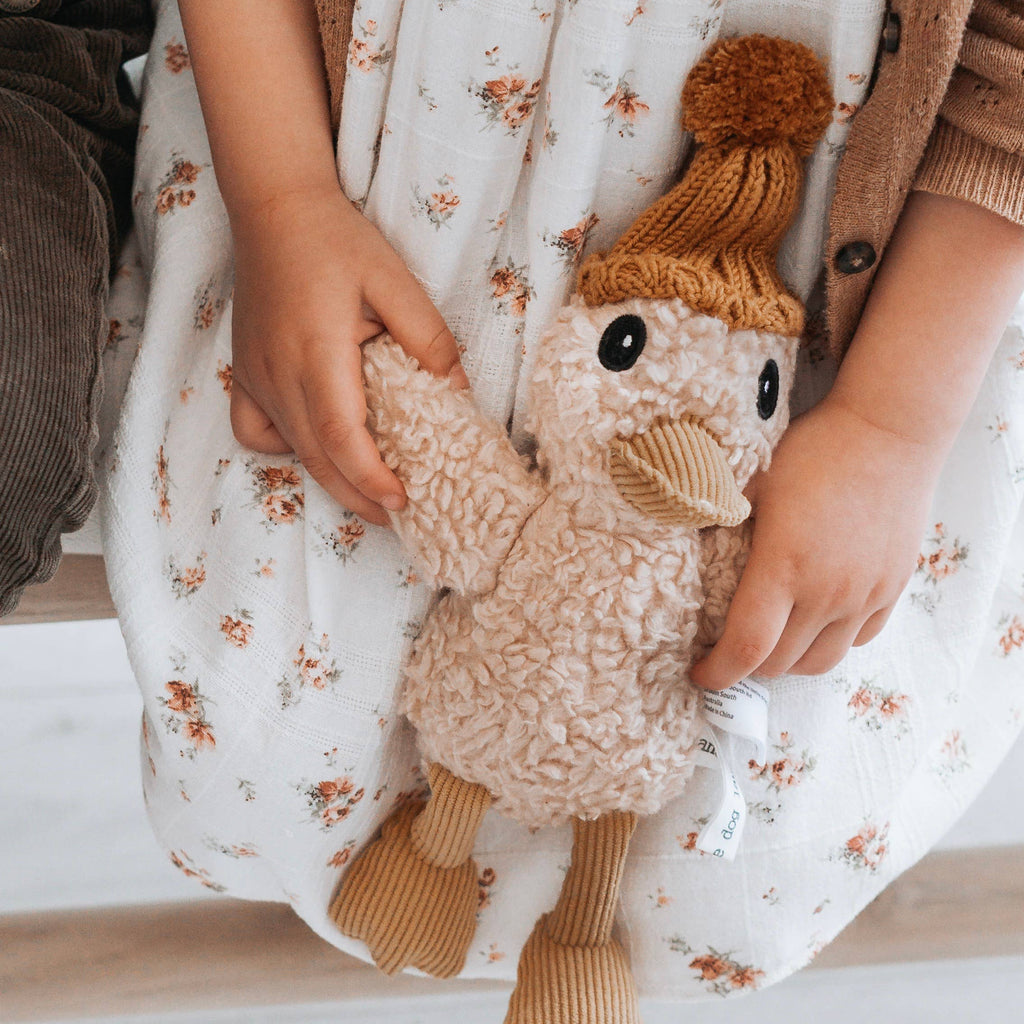 Child holding Petunia Duck soft toy made from beige cotton plush with caramel corduroy beak and feet, wearing a brown merino wool knitted beanie.