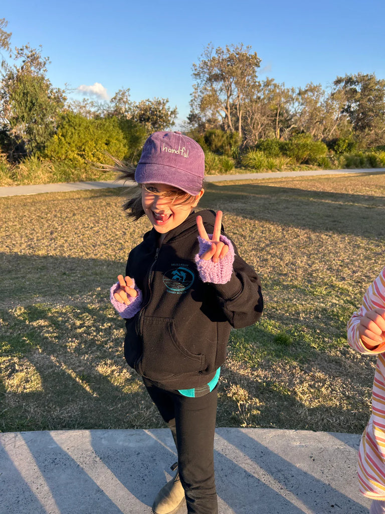 Young child wearing the purple "Handful" kids cap in a sunny outdoor setting, highlighting the playful spirit and comfortable fit of this fun accessory.