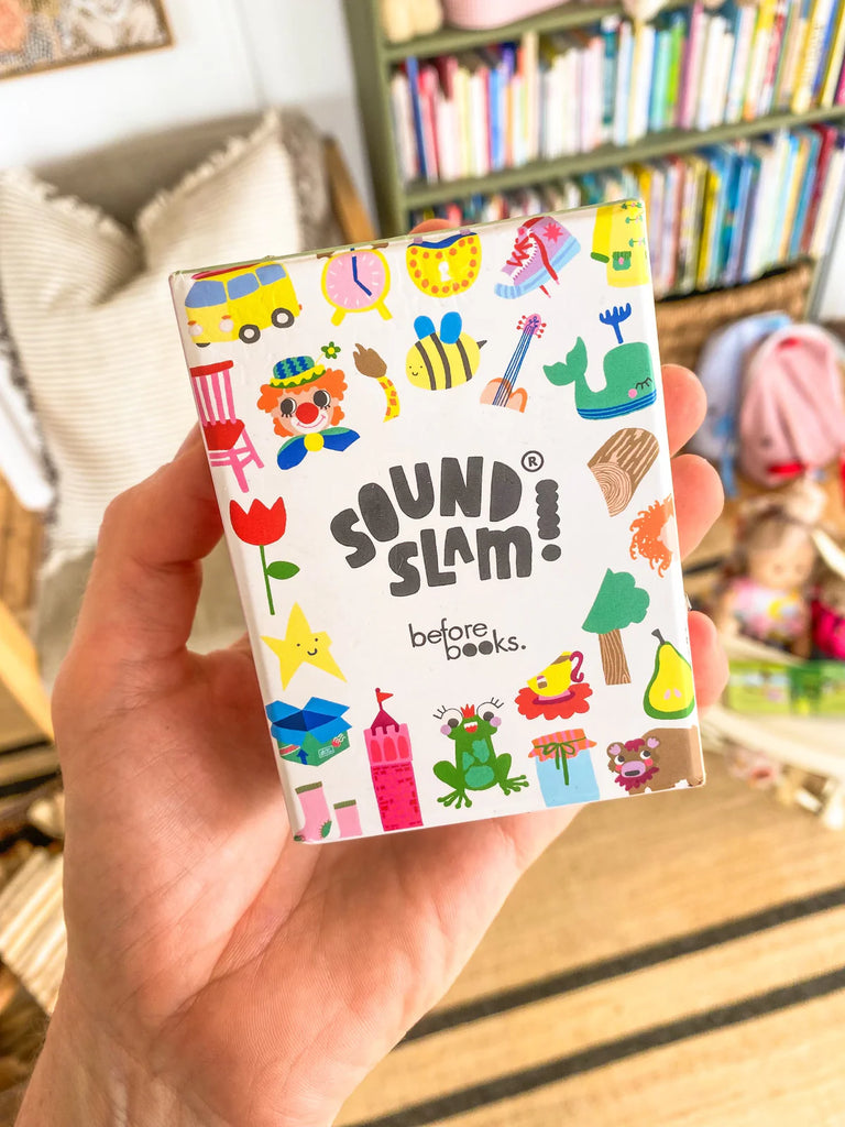 Hand holding the Sound Slam card game box, featuring bright, playful illustrations of objects and animals designed to support phonological awareness for children. Bookshelf and toys visible in the background, adding a warm, educational setting.