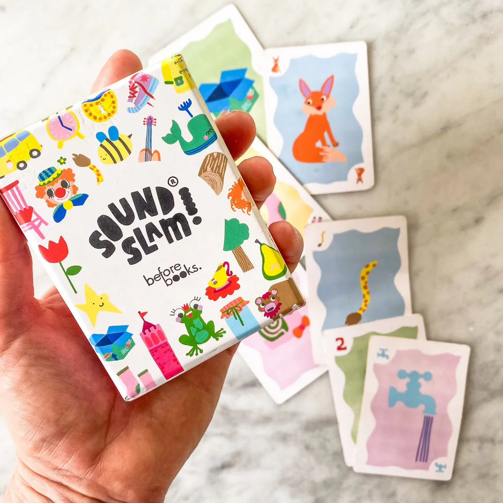 Hand holding the colourful Sound Slam game box with illustrated cards scattered on a marble table, showing the vibrant, letter-free learning design.