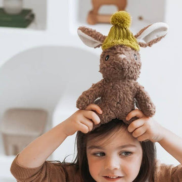 Child holding Stella Rabbit soft toy on their head, showing brown cotton plush fur, floppy ears, and mustard merino wool beanie.