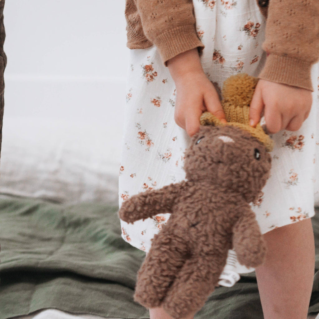 Close-up of child’s hands holding Stella Rabbit plush toy, highlighting caramel brown plush fabric and mustard knitted beanie.