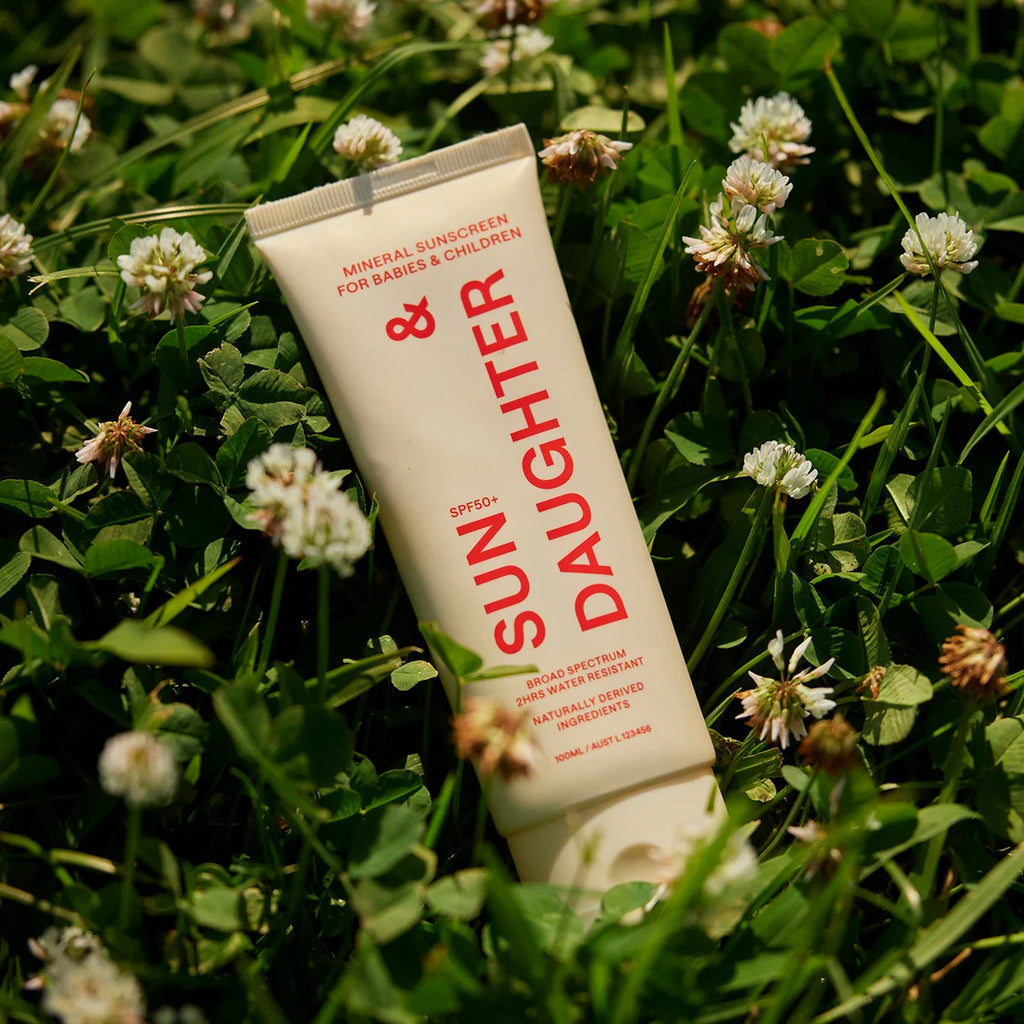 Sun & Daughter SPF50+ Mineral Sunscreen tube lying in green grass surrounded by white clover flowers.