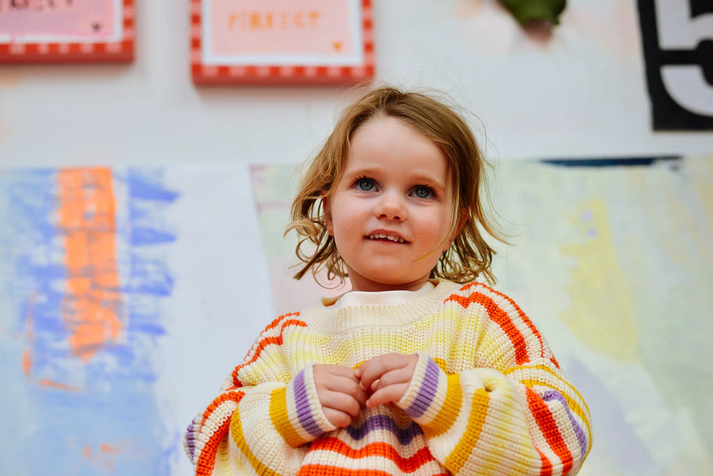 Young child holding up the Sunday Siblings Art Club Knit in front of an abstract, pastel wall mural, showcasing the bold stripe pattern and relaxed fit.