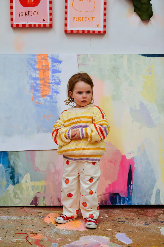 Child in the Sunday Siblings Art Club Knit smiling brightly, styled in playful retro fashion with pastel pants, standing on a paint-splattered floor.