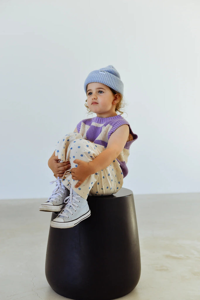 Young child sitting on a black ottoman wearing the Blue Figo Beanie, purple top, and printed pants, looking off to the side in a clean, minimal backdrop.