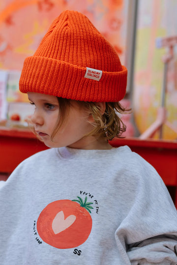 Side profile of a child wearing the bright red Figo Beanie by Sunday Siblings, styled with a grey tomato-print sweatshirt in a playful, pastel art space.