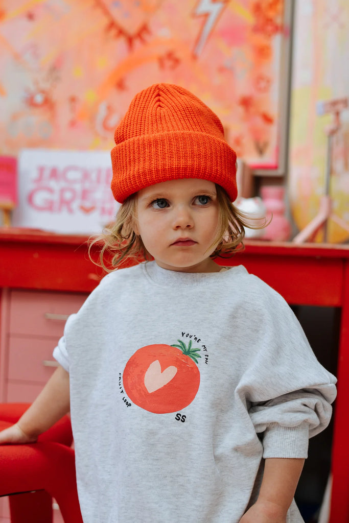 Toddler in a vibrant red Figo Beanie and oversized grey top with tomato graphic, standing confidently in front of a colourful studio setup.