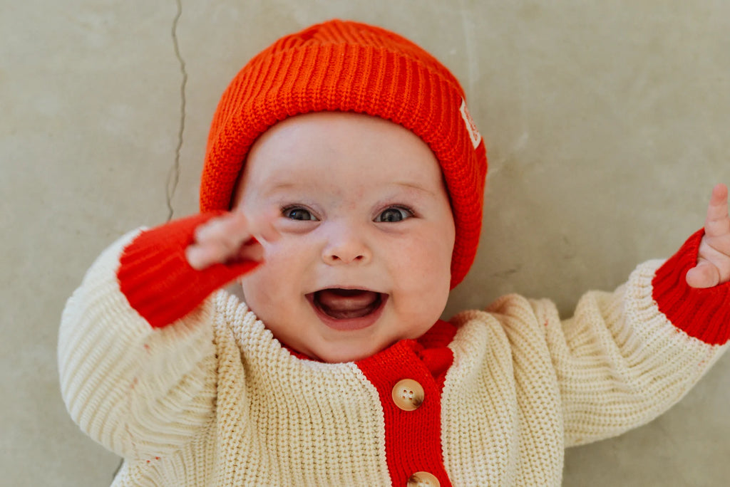 Smiling baby in the red Figo Beanie and cream cardigan with red accents, reaching up joyfully in a soft, neutral-toned background.