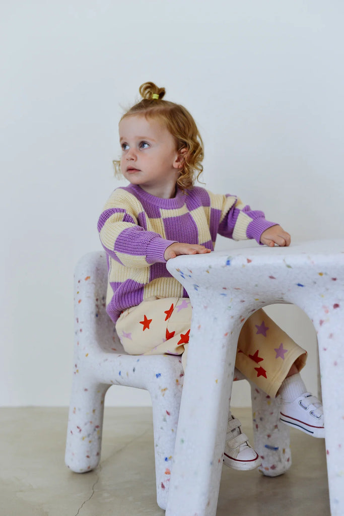 Child wearing the purple Sunday Knit jumper, sitting and smiling in a playful pose, styled with pastel patterned pants in a minimalist indoor setting.