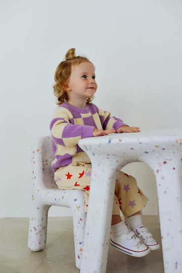 Toddler sitting on a white speckled chair, wearing a purple and cream checkered knit jumper by Sunday Siblings, paired with printed pants and white sneakers.