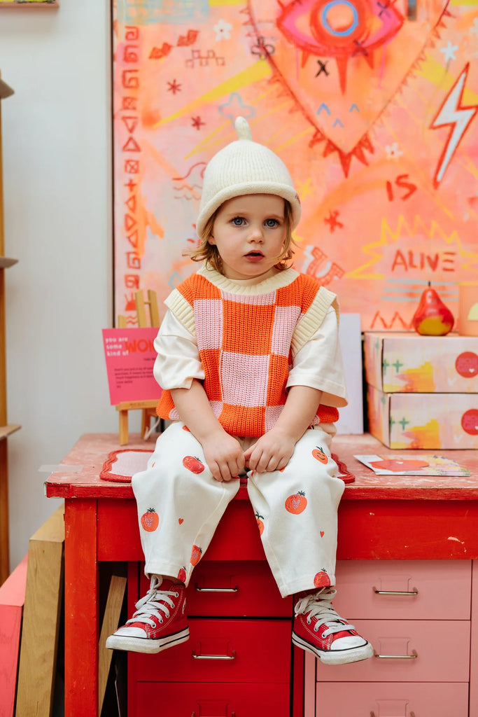 Toddler wearing the cream Teepee Beanie by Sunday Siblings, seated on a red desk with feet dangling, styled in colourful retro-inspired kidswear.