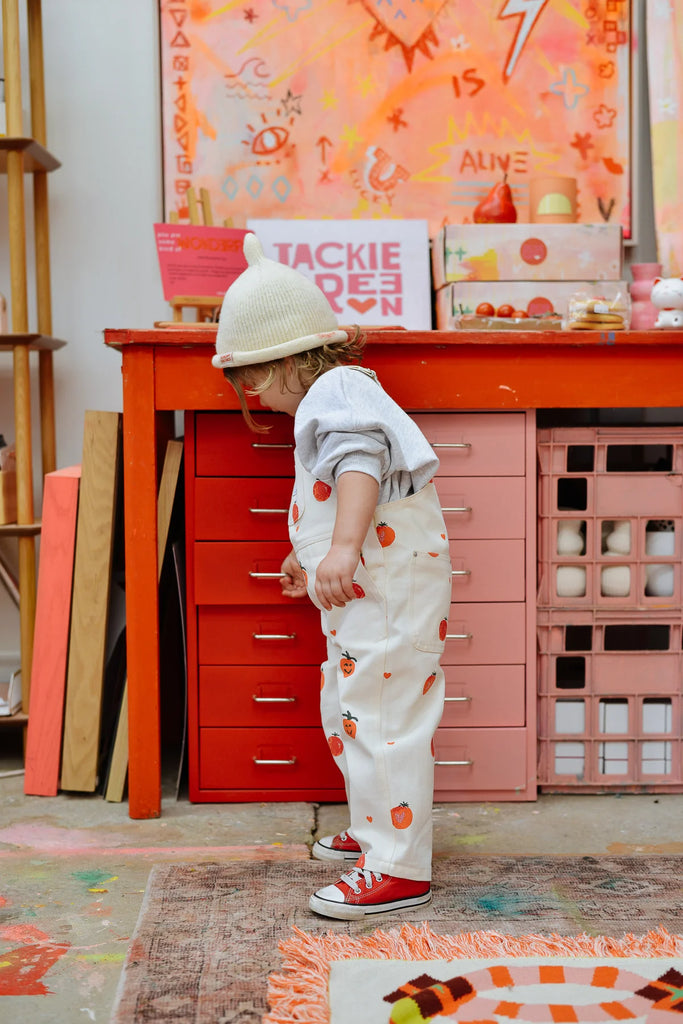 Side profile of a toddler wearing a cream Teepee Beanie, grey tee, and printed pants, leaning on a red desk surrounded by playful, creative decor.