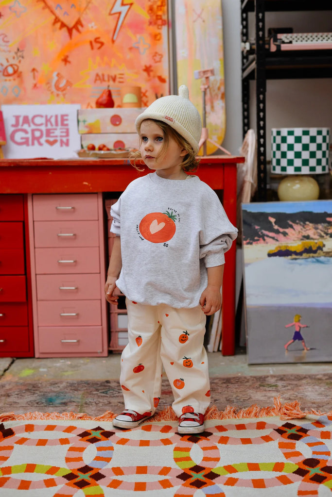 Young child in a cream Teepee Beanie, oversized grey tee and heart-print pants, standing on a vibrant patterned rug in an art-inspired studio space.