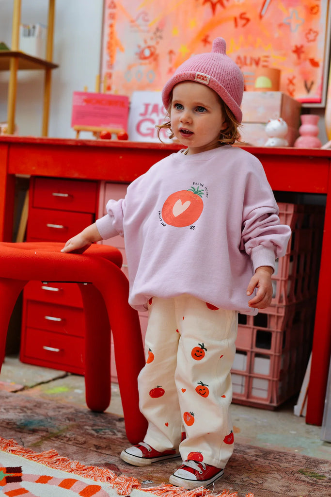 Toddler side profile in a pink Teepee Beanie and fun printed pants, posing in a colourful studio space with art-inspired decor and bold red furniture.