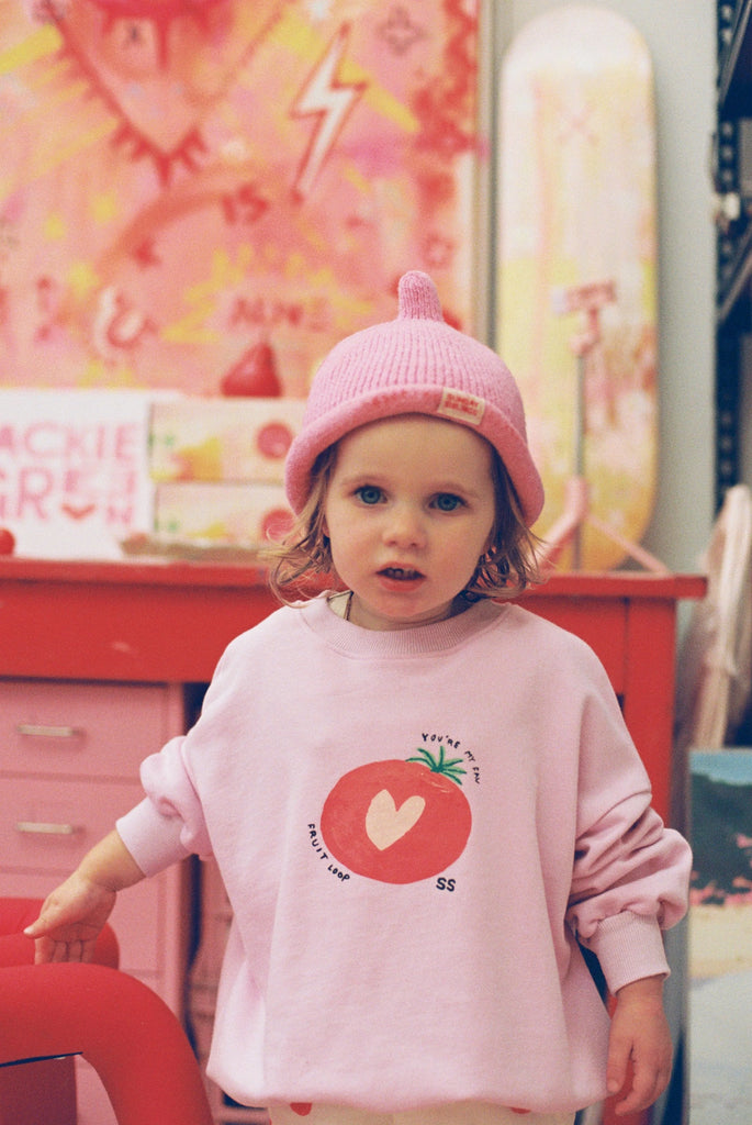 Young child in a pink Teepee Beanie and oversized blush top with orange graphic, standing in front of a red art table, smiling in a vibrant, playful setting.