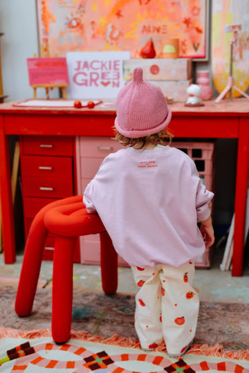 Toddler wearing a pink Teepee Beanie and pastel outfit with red and white patterned pants, standing at a bright red art desk in a colourful playroom.