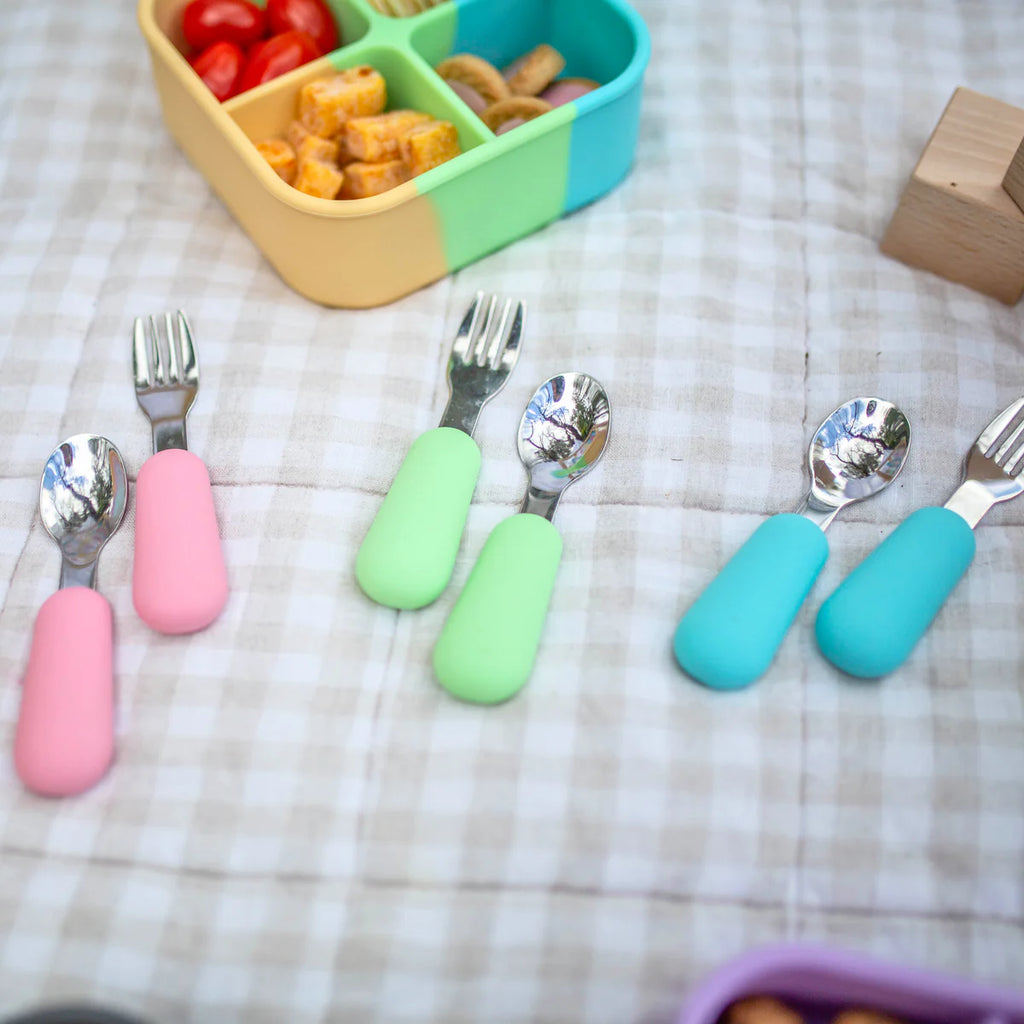 Close-up of 3 colourful spoon and fork sets next to lunchboxes filled with snacks, set on a beige gingham picnic rug.