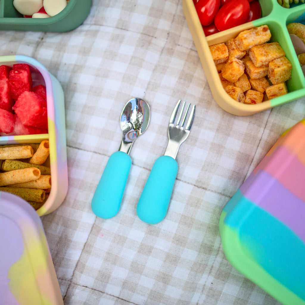 Close-up of aqua spoon and fork set beside fruit-filled lunchboxes on a picnic rug.