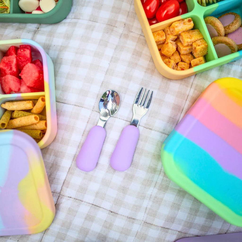 Close-up of lilac spoon and fork set beside fruit-filled lunchboxes on a picnic rug.