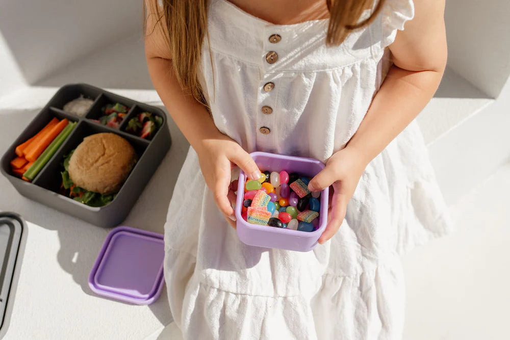 Toddler in a white dress holding a lilac Zero Waste People snack container filled with colourful food items, with a black bento lunchbox nearby.