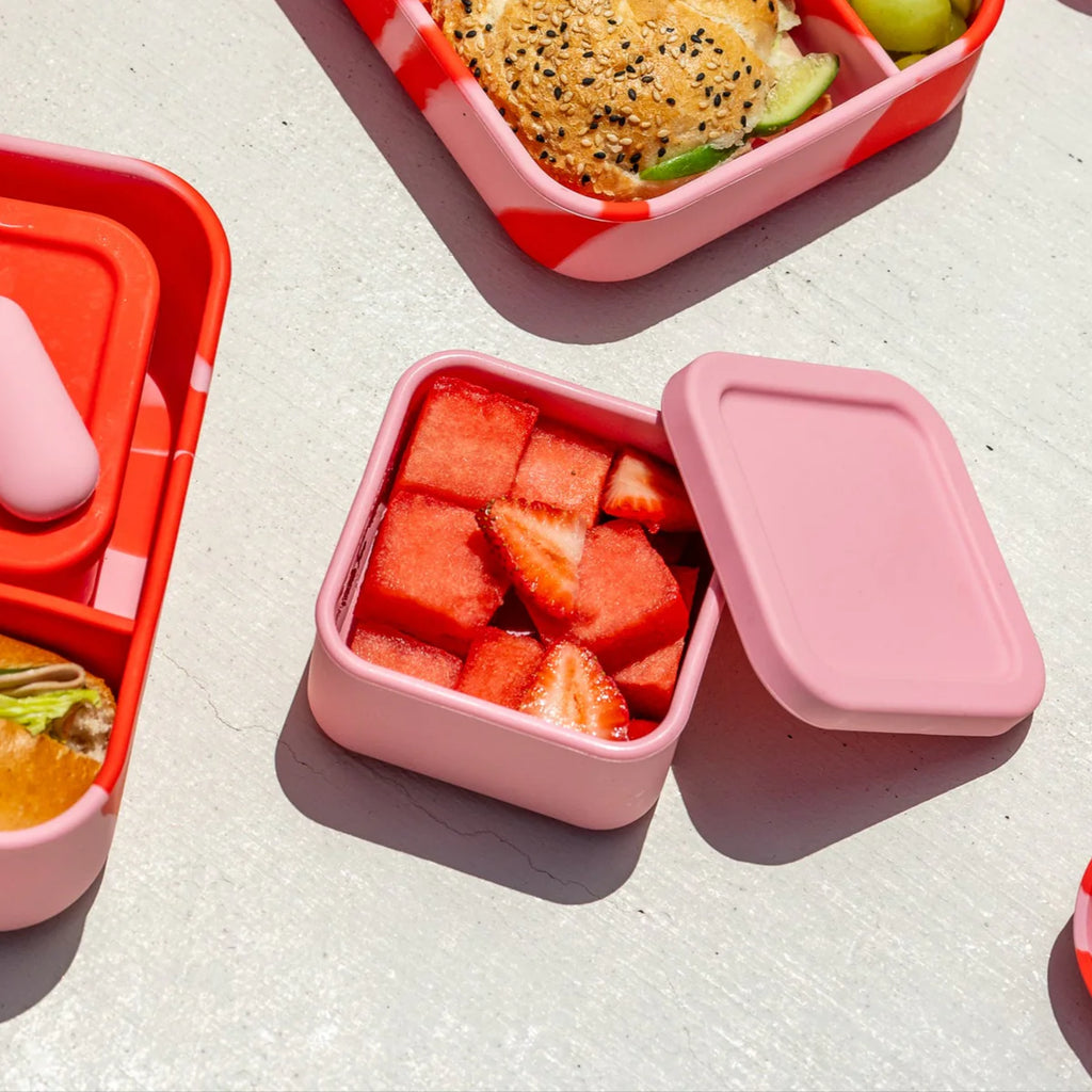 Close-up of The Zero Waste People snack container in watermelon pink, filled with fresh strawberries and watermelon, next to a red swirl bento lunchbox.