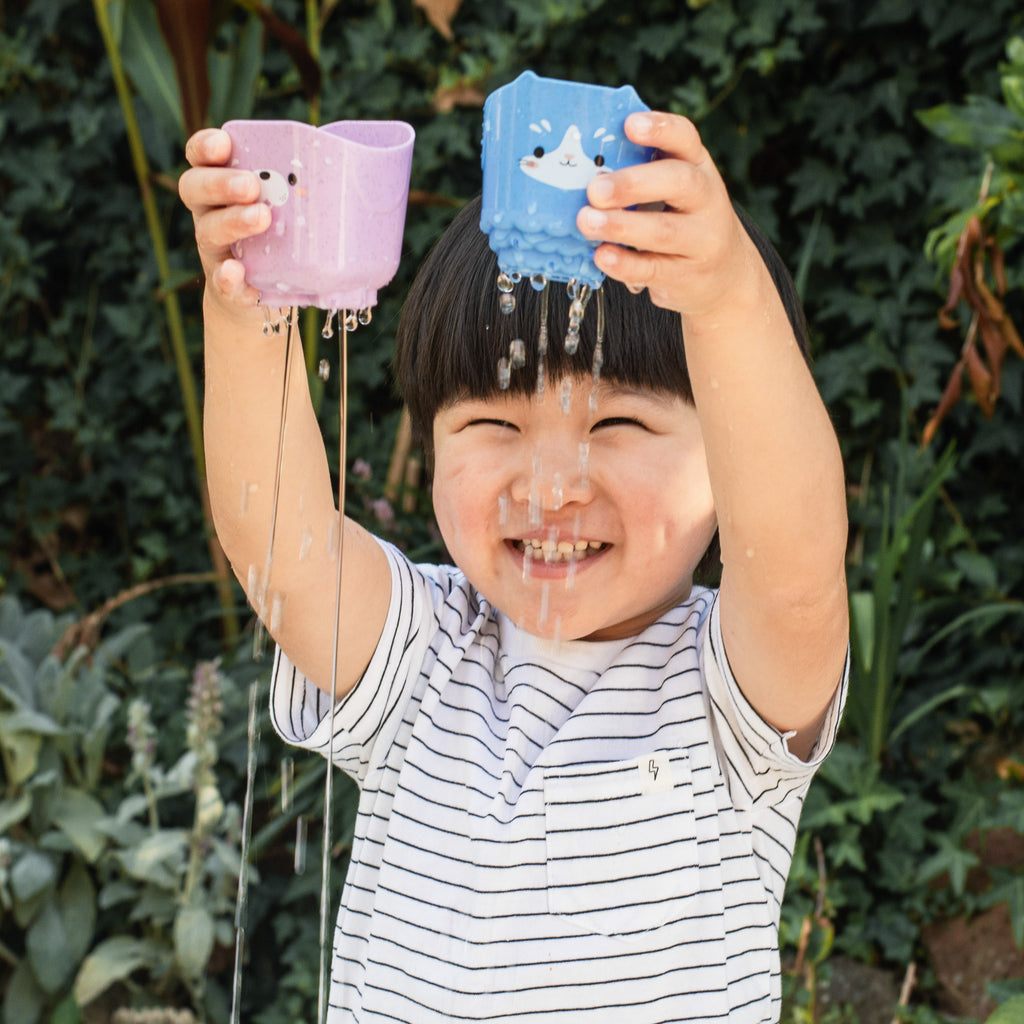 Young child lifting and pouring from the animal cups, showing joyful engagement and active bath-time-style fun in a garden or water area.