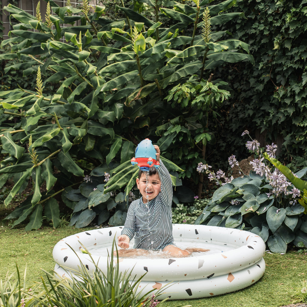 Child using the Bath Rocket in an inflatable pool outdoors, smiling and lifting it overhead as water pours down, engaging in sensory splash play.