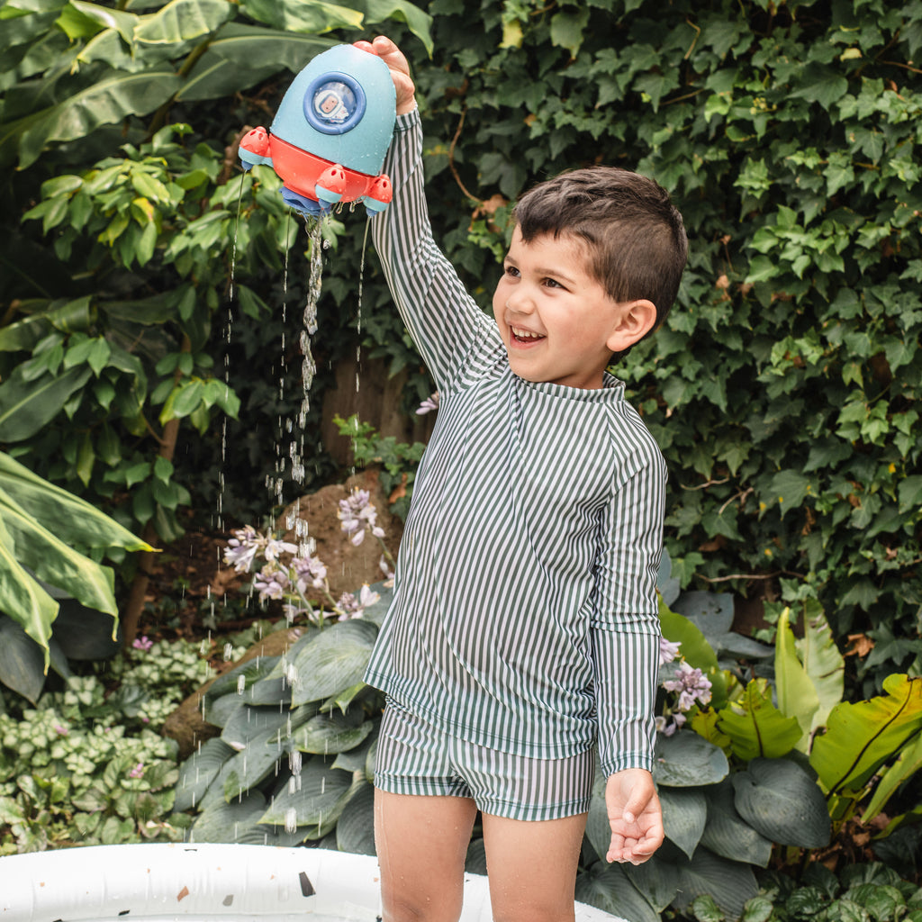 Toddler boy raising the Bath Rocket and watching water pour out, showing developmental motor engagement and enjoyment during water play.