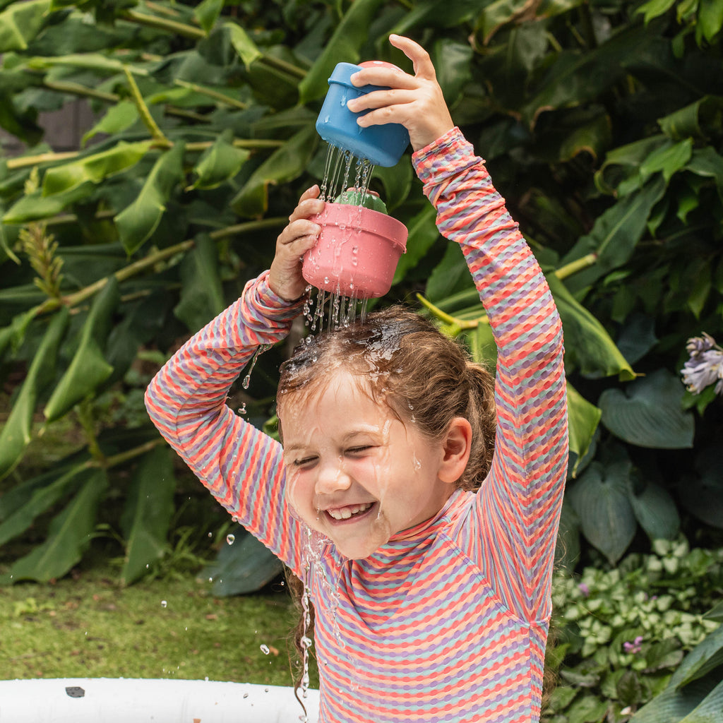 Young child laughing and tipping the cactus bath toy overhead outdoors, enjoying the water sprinkling feature and sensory surprise.