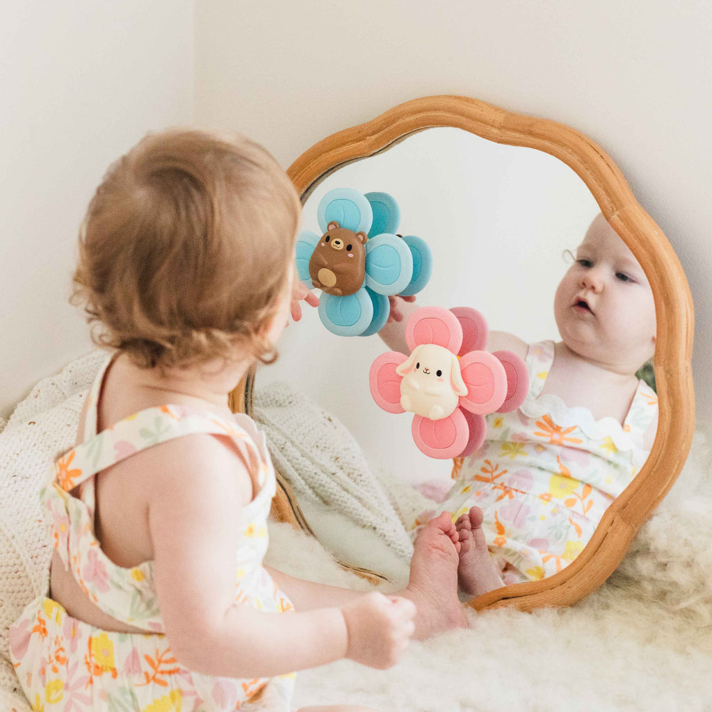 Baby sitting in front of a wooden-framed mirror, engaging with two colourful sensory spinners—one blue with a bear and one pink with a bunny—both suctioned to the mirror, encouraging fine motor play and visual exploration.