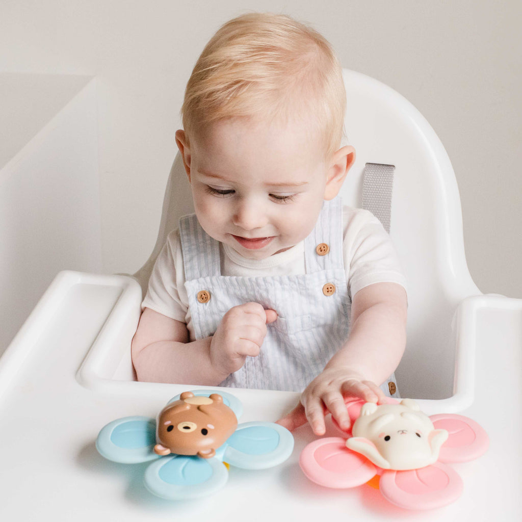 Baby sitting in a white high chair spinning both the Bunny and Bear sensory toys, showing their easy suction attachment and play value during mealtimes.